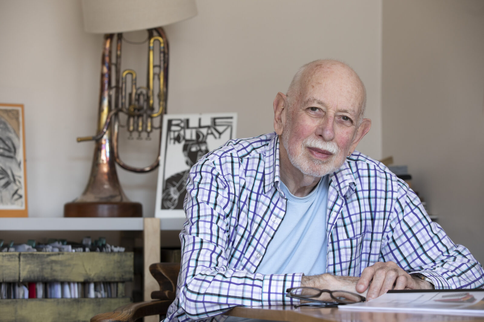 An older man sitting at the table in his living room, smiling, with a brass instrument and a Billie Holiday poster behind him