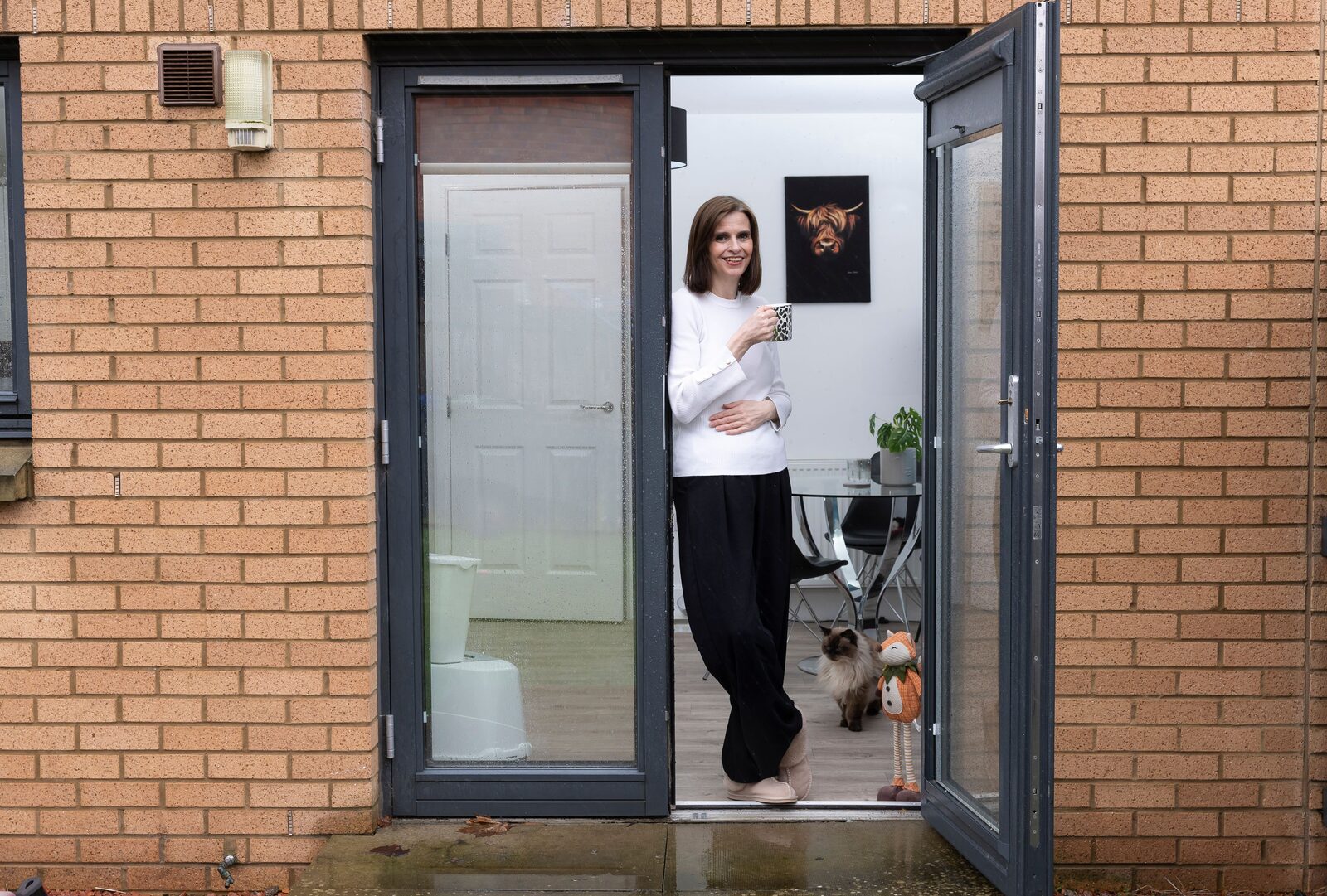 A woman standing in the open back door of her flat holding a mug, with her cat behind her