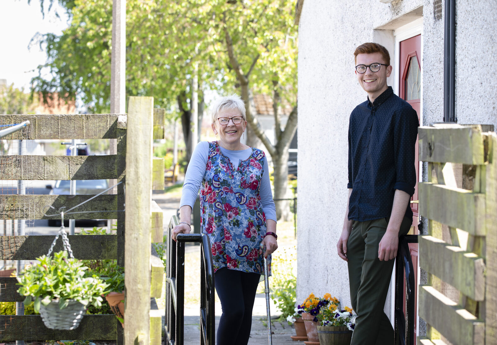 An older woman with a walking stick at her garden gate, smiling, with a young man standing by her front door